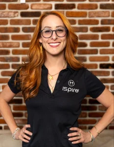 A woman with long red hair and glasses smiles while standing in front of a brick wall. She is wearing a black polo shirt with the "Ispire" logo, along with various necklaces and bracelets. With her hands on her hips, she looks ready to share Vaporizer Resources and testimonials alike. CA Norml