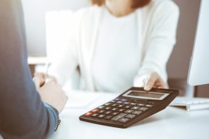 A person in a white long-sleeve shirt handing a calculator to another individual whose back is turned, while sitting at a desk with business documents and a computer monitor. CA Norml