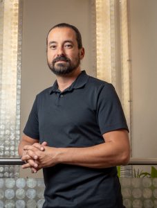 A man with a beard and mustache is standing in front of a textured wall, wearing a black polo shirt. His hair is tied back, and he has his hands clasped together in front of him. With a calm and composed expression, he looks like one of those trusted doctors giving testimonials about their practice. CA Norml