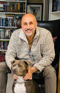 A bald man with a short beard, wearing a fluffy grey sweater and glasses hanging from his neck, is seated and smiling. He has both hands affectionately on a large brown and white dog sitting in front of him. A bookshelf filled with books is in the background, showcasing testimonials from satisfied retailers. CA Norml