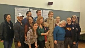 A group of thirteen adults and one child from the ReformCA Coalition pose together in a classroom, smiling. Some stand while others crouch in front. Behind them are handwritten notes on large paper sheets taped to a chalkboard. CA Norml