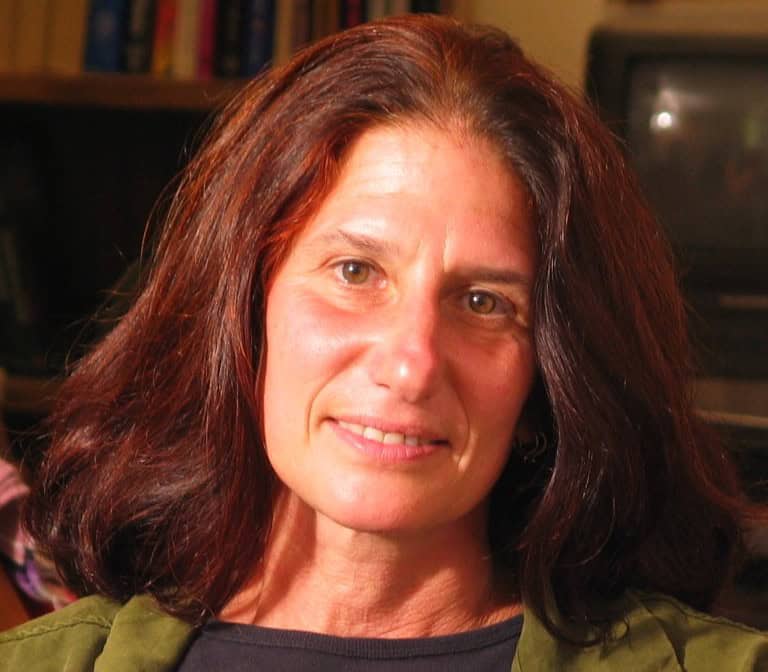 A person with shoulder-length brown hair and a warm smile is looking at the camera. They are wearing a dark shirt and a green jacket. The background is blurred but contains some shelves with books, hinting at their involvement in the Cal NORML Board of Directors, and a television screen. CA Norml