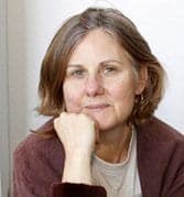 A woman with shoulder-length brown hair, wearing a brown cardigan and a beige top, rests her chin on her hand while looking at the camera with a neutral expression. She is seated against a light-colored background, embodying the calm demeanor of someone on the Cal NORML Board of Directors. CA Norml