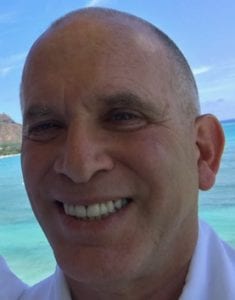 A close-up photo of a man smiling at the camera with a scenic background of the ocean and blue sky. He is wearing a white shirt and appears to be outdoors, embodying the relaxed spirit of Cal NORML's Board of Directors. CA Norml