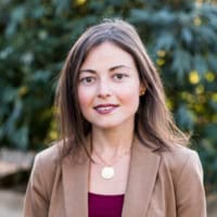 A woman with long brown hair is standing outside, dressed in a tan blazer over a burgundy top. She is smiling slightly and looking directly at the camera. The background features green foliage, adding an earthy touch to her composed demeanor as a member of the Cal NORML Board of Directors. CA Norml