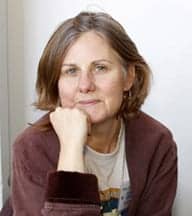 A person with shoulder-length brown hair sits resting their chin on their hand, wearing a brown cardigan over a light-colored shirt and looking directly at the camera. The background is a plain white wall, reminiscent of the promotional photos taken for the Cannabis exhibit at the CA State Fair on July 31. CA Norml