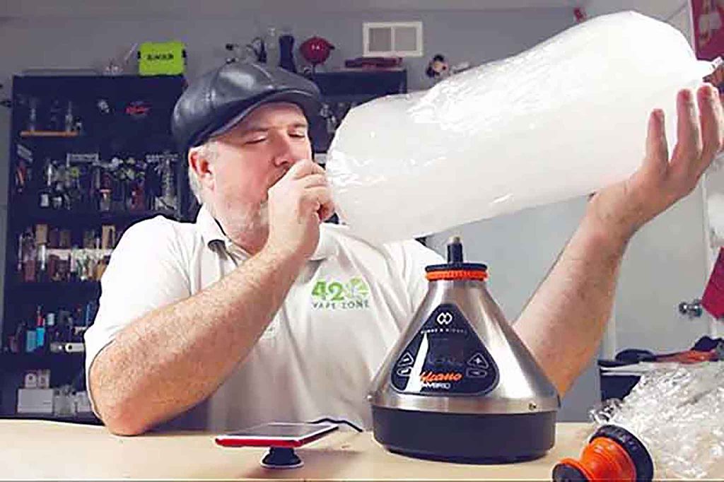 A man in a white shirt and black cap inhales from a large, transparent vapor-filled bag attached to a tabletop vaporizer on a cluttered counter, enjoying his session in the 420 Vape Zone. CA Norml