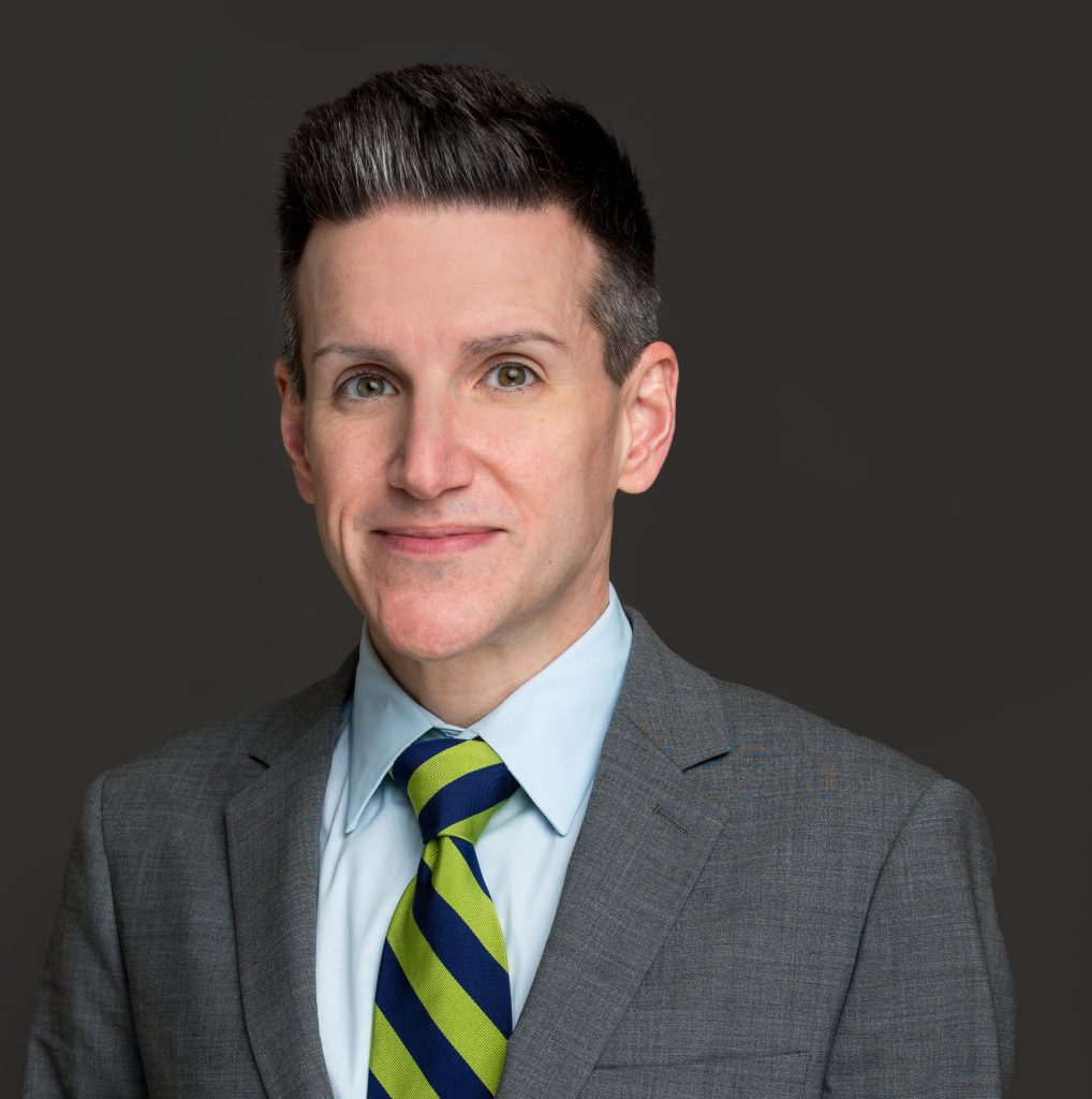 Shay Aaron Gilmore, a man with short dark hair, wears a gray suit, light blue shirt, and green and navy striped tie while posing in front of a plain dark background. CA Norml