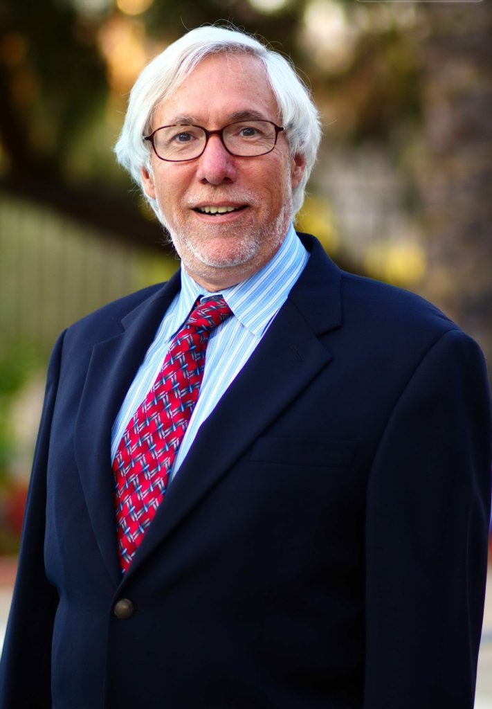 An older man, S. Edward Wicker, with white hair and glasses is smiling outdoors. Dressed in a navy suit, blue-striped shirt, and red patterned tie, the attorney stands confidently amidst a backdrop of trees and greenery. CA Norml