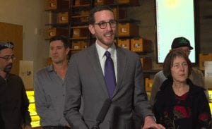 A man in a gray suit and purple tie stands behind a microphone, speaking with compassion to an audience. He is positioned in front of several people. Shelves with boxes are visible in the background, and one person in the background is wearing a cap with a cannabis leaf symbol. CA Norml