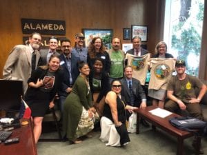 A group of people posing together in an office room labeled Alameda, featuring wooden walls, framed pictures, and a window. Some individuals are seated while others are standing, and two people are holding up graphic t-shirts from ASA's Lobby Day as part of the Cal NORML initiative. CA Norml