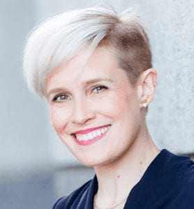 A person with short, platinum blonde hair and a side undercut is smiling. They are wearing a dark-colored top and small, stud earrings. The background appears to be a light gray wall, reminiscent of the setting for the California NORML Legal Seminar in June 2021. CA Norml