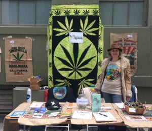 A person stands behind a table with various items, including papers, pamphlets, and paraphernalia related to marijuana. A large banner with a marijuana leaf is displayed behind them. Representing Cal NORML, they proudly wear a hat and t-shirt with a marijuana-themed design while sharing 2018 accomplishments and plans for 2019. CA Norml