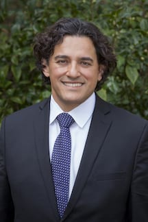 A person with wavy hair, wearing a dark suit, white shirt, and patterned tie, smiles at the camera. The background is blurred with green foliage. CA Norml