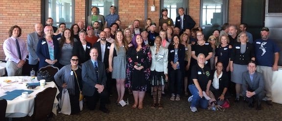 A diverse group of around 50 people posing for a group photo in a conference room with brick walls in Sacramento. Some are standing, others are sitting at tables. They appear to be dressed in business casual attire and are smiling towards the camera during their Annual Lobby Day on May 6. CA Norml