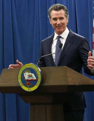 A man stands at a podium with a seal that says "Governor of the State of California." He is wearing a dark suit and tie, speaking into a microphone with his hands gesturing. Discussing statutory changes, the background is a blue curtain, and an American flag is partially visible on the side. CA Norml