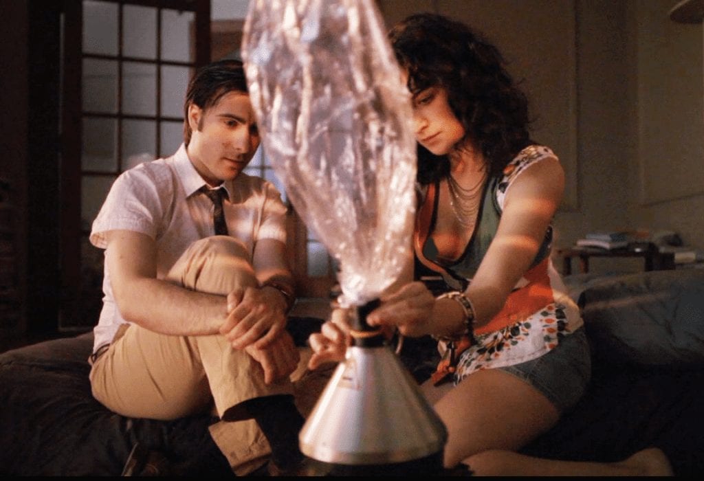 Two people are sitting on the floor. The person on the left is wearing a white shirt and light-colored pants, while the person on the right, dressed in a patterned top and denim shorts, is handling a large silver object with a transparent bag attached. The room is dimly lit; it's clear this setup isn't safe for cannabis vaping indoors. CA Norml
