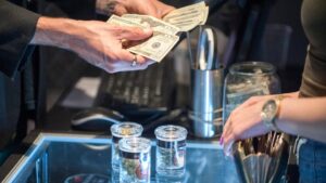 A close-up of two people exchanging cash on a glass display counter. One person is handing over dollar bills, while the other person's hand, wearing a gold watch, is poised near a transparent jar filled with more dollar bills. Three small glass containers are visible on the counter, perhaps for cannabis products endorsed by NORML. CA Norml
