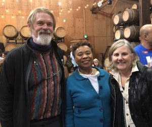 Three people are standing and smiling at a gathering in a rustic, wood-paneled setting with barrels stacked in the background. The group consists of a man with a beard wearing a sweater and jacket, a woman in a blue jacket, and another woman in a black jacket, likely discussing their 2019 accomplishments. CA Norml