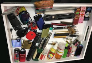 A drawer filled with various smoking and vaping devices, e-liquid bottles, lighters, and accessories. The items are assorted, including vape pens, cartridges, and colorful containers from Cal NORML's 2019 accomplishments, all arranged in a somewhat cluttered manner. CA Norml