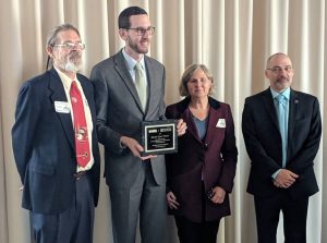 A group of four people stand together, three men and one woman, dressed in formal attire. The man second from the left holds a rectangular plaque celebrating 2019 Accomplishments and is smiling. They are posing in front of a light-colored curtain. CA Norml