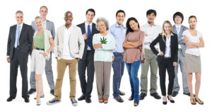 A diverse group of 12 adults stand in a row, facing the camera. They are dressed in a variety of outfits ranging from casual to business attire. One woman is holding a cannabis leaf symbol on her shirt, representing Cal NORML's 2019 accomplishments. The background is plain white. CA Norml