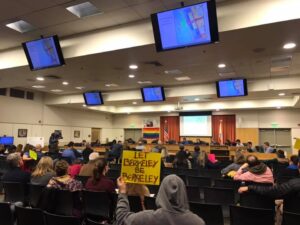 A group of people are seated in a meeting room, facing a panel of officials discussing cannabis licensing. Multiple screens display maps, and a rainbow flag is hung on the wall. One attendee holds a yellow sign saying "LET BERKELEY BE BERKELEY. CA Norml
