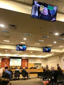 A community meeting is taking place in a large room with people seated and facing a panel of speakers. Several screens display a person speaking, while protest signs in the background read "BAN'S DON'T WORK" and "WE ARE ALL WIRE SMALL." The room, possibly in Berkeley, has high ceilings and bright lighting. CA Norml