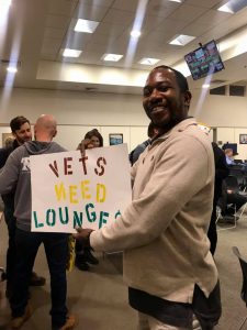 A person smiles while holding a sign that reads "Vets Weed Lounges" in a public room in Berkeley. Other individuals are in the background, engaging in conversation about cannabis licensing. The room has a high ceiling with lights and a TV screen mounted on the wall. CA Norml