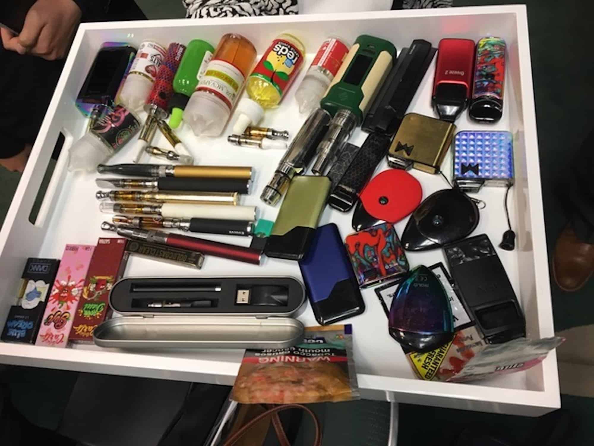 A white tray is filled with various vaping supplies, including multiple vape pens, e-cigarettes, vape mods, cartridges, USB chargers, and bottles of flavored e-liquid. Flavored e-liquid packaging and a range of colorful and metallic devices are visible. For more insights on vaping trends, see the latest Cal NORML white paper. CA Norml