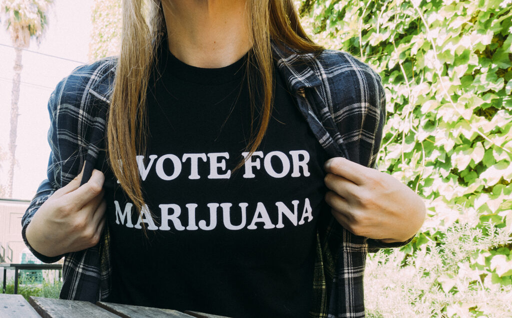 A person with long hair wearing a black t-shirt with the text "VOTE FOR MARIJUANA" printed on it holds open their plaid shirt to display the message. Against a backdrop of green foliage and sunlight, they emphasize support for CA Special Elections on May 12. CA Norml