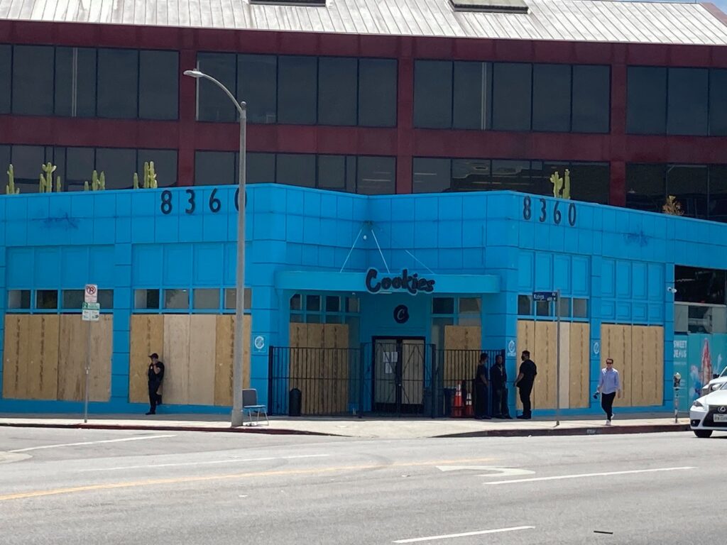 A building with a bright blue exterior stands on a street corner. The word "Cookies" is written above the entrance, and the windows and doors are covered with wooden boards, hinting at recent closures. Several people are standing or walking near the building. The address number "8360" is visible. CA Norml