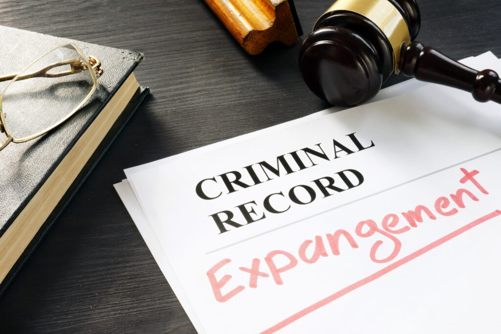 A document labeled "CRIMINAL RECORD" with the word "Expungement" written in red sits on a dark desk. Nearby are a pair of glasses, a closed book, and a judge's gavel. The scene signifies steps taken in California for clearing past marijuana crimes from individual records. CA Norml