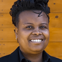 A person with short curly hair and a broad smile, wearing a black blazer and black shirt, poses in front of an orange wooden background, symbolizing the vibrant spirit of racial justice. CA Norml