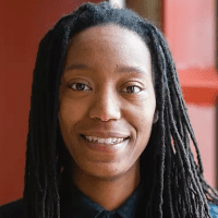 A person with long dreadlocks wearing a black shirt smiles while facing the camera. The background features red tones, suggesting an indoor setting with natural light, creating an atmosphere that resonates strongly with themes of racial justice and equity. CA Norml