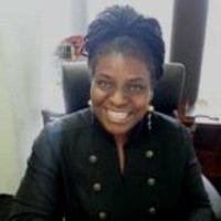 A person with braided hair is sitting and smiling at the camera. They are wearing a dark jacket with a buttoned front, and there are two large windows with light curtains behind them. The setting appears to be an office focused on equity and racial justice. CA Norml