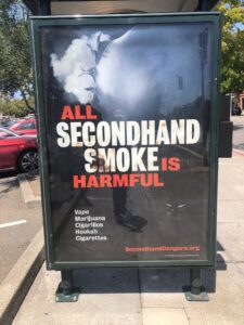A bus stop advertisement shows large text reading, "ALL SECONDHAND SMOKE IS HARMFUL," with "HARMFUL" in red. Below, smaller text lists "Vape Hazards, Marijuana, Cigarillos, Hookah, Cigarettes." The background features smoke, and the website "SecondhandDangers.org" appears at the bottom. CA Norml