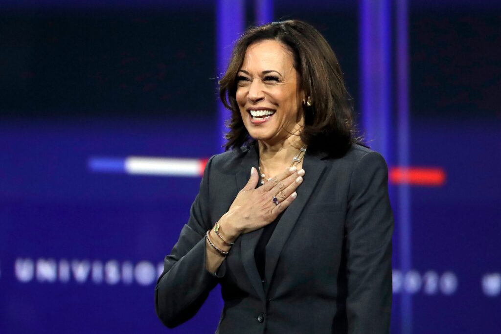 A woman in a dark blazer stands on stage, smiling and holding her hand to her chest. The background features blurred, dark blue elements and indistinct text. She appears to be speaking or acknowledging an audience, reminiscent of Kamala Harris when she was introduced as Biden's running mate. CA Norml