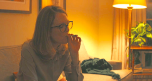 A person with long hair and glasses is sitting on a sofa in a dimly lit room, holding a lit cigarette to their mouth. The background features a warm, yellowish table lamp, a potted plant, and partially curtained windows, evoking an atmosphere evocative of discussions around the San Francisco Ban on Cannabis Smoking. CA Norml