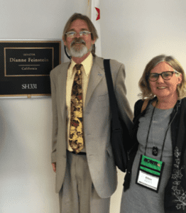 Two people standing indoors in front of a sign that reads "Senator Dianne Feinstein, California, SH331." The man on the left has gray hair, a beard, glasses, a beige suit, and a patterned tie. The woman on the right has gray hair, glasses, and is wearing a black jacket and name badge from Cal NORML. CA Norml