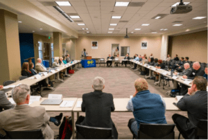 A large conference room with people seated around three sides of a U-shaped arrangement of tables. A speaker stands at a podium on the left, discussing the latest Impaired Driving report, while other attendees listen attentively. A blue banner with yellow letters is displayed at the front of the room. CA Norml