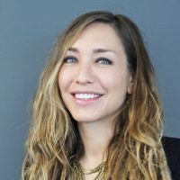 A woman with long, wavy blonde hair and blue eyes is smiling. She is wearing a dark top with a layered gold necklace. The background, reminiscent of the California NORML seminar in June 2021, is a solid gray color. CA Norml