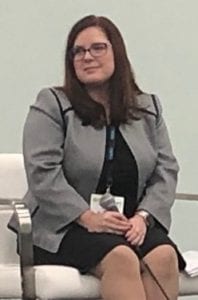A woman with long brown hair and glasses, wearing a gray blazer over a black dress, sits on a white chair. She holds a microphone and is looking to the side, likely preparing for her speech at the Legal Seminar hosted by California NORML on June 1 & 2. CA Norml