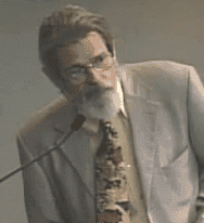 A person with glasses, a mustache, and a beard is speaking into a microphone. They are wearing a light-colored suit with a patterned tie. The plain grey background suggests an indoor setting, possibly during the California NORML Legal Seminar in June 2021. CA Norml