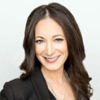 A woman with long, dark hair and blue eyes is smiling at the camera. She is wearing a black blazer and a necklace with a small pendant. The background is plain and light-colored, reminiscent of the setting for the California NORML Legal Seminar in June 2021. CA Norml