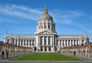 A grand neoclassical building with a large central dome and rows of symmetrical columns stands against a clear blue sky. In front, a manicured lawn is bordered by paths and trees, with a few people walking along them. Flags adorn the building, which occasionally hosts Oakland City Council meetings. CA Norml