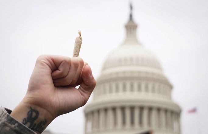 A hand holding a rolled joint is raised in the foreground, with the United States Capitol building in the background. The hand has a visible tattoo near the wrist, and the sky is overcast. CA Norml