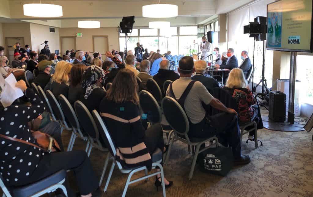 A diverse group of people attends an indoor conference on Cal NORML's 2021/2022 activities. They are seated facing a panel of speakers. A large screen displays a presentation. Some individuals are wearing masks. The room is well-lit with natural light streaming through large windows. CA Norml