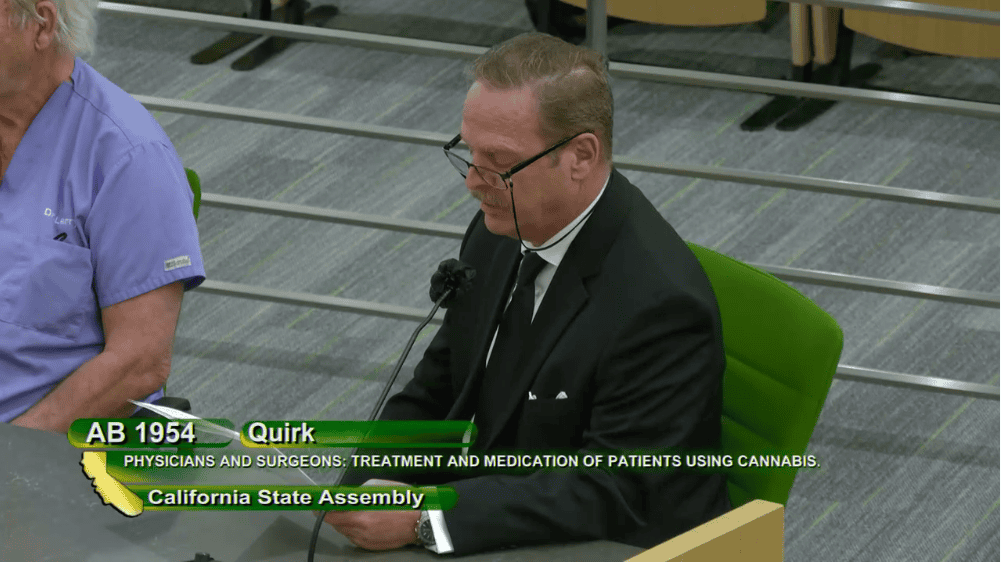 A man in a black suit and tie, wearing glasses, speaks into a microphone at a California State Assembly hearing. A person in scrubs is seated next to him. The screen text reads: "AB 1954 Quirk - Physicians and Surgeons: Treatment and Medication of Patients Using Cannabis," emphasizing medical rights for cannabis users' employment. CA Norml