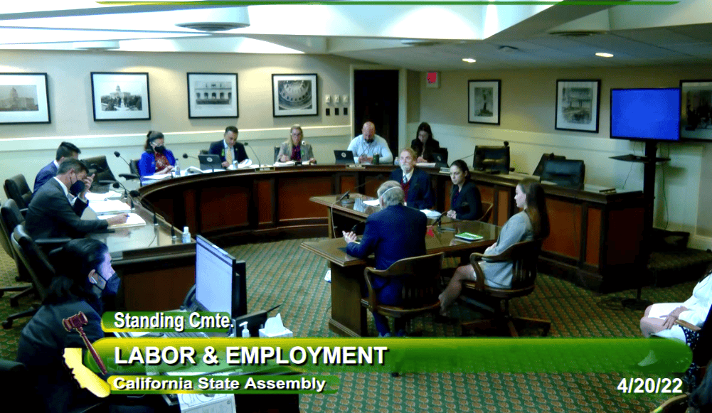 A meeting of the Standing Committee on Labor & Employment in the California State Assembly is in session. Several people are seated around a table, engaged in discussing employment rights. A display screen and various office equipment are visible in the room. The date is 4/20/22. CA Norml